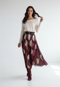 Kremowa bluzka z odkrytymi ramionami zestawiona z plisowaną bordową spódnicą w wyraziste okrągłe wzory, przewiązana czarnym paskiem, do tego bordowe kozaki za kolano.