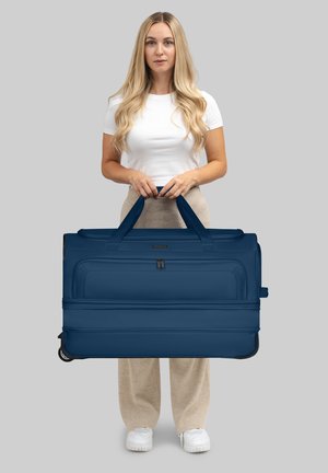 Una donna con capelli biondi lunghi, che indossa una camicia bianca e pantaloni beige, tiene una grande valigia blu navy con rotelle per le maniglie.
