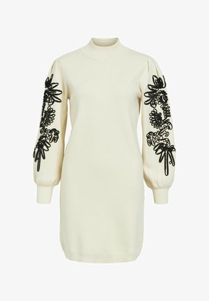 Vestido de punto crema con un cuello alto, mangas abullonadas y bordado floral negro a lo largo de las mangas. Textura suave y forma ajustada.