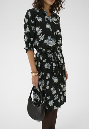 Schwarzes Blumenkleid mit blauen und weißen Mustern, Knopfleiste vorne, hochgekrempelte Ärmel und Taillengürtel. Hält eine schwarze Halbmond-Handtasche, trägt ein goldenes Armband.