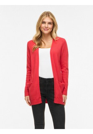 Femme aux longs cheveux blonds portant un cardigan rouge ouvert sur un haut blanc et un jean noir, debout devant un fond blanc.