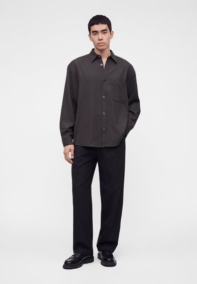 Homme debout portant une chemise ample et foncée à boutons, un pantalon noir à jambes larges et des chaussures noires sur un fond clair uni.