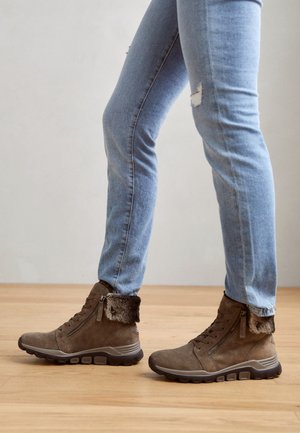 Person iført lyseblå, revnede jeans og brune ankelstøvler foret med pels, stående på trægulv.