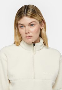 Pullover in pile con zip di colore crema, caratterizzato da una superficie testurizzata. Presenta un colletto alto e un ricamo logo discreto sul petto.