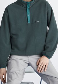 Mørkegrøn fleecepullover med en teal knapkrave, der har en brystlomme og to sidelommer samt elastiske ærmer for en tæt pasform.