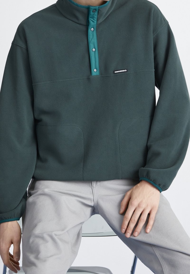 Mørkegrøn fleecepullover med en teal knapkrave, der har en brystlomme og to sidelommer samt elastiske ærmer for en tæt pasform.
