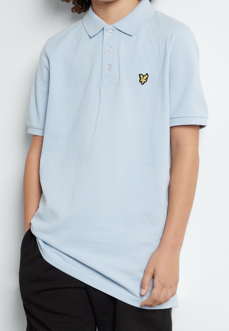 Enfant portant un polo à manches courtes bleu clair avec un logo d'oiseau jaune sur la poitrine et un pantalon noir, debout avec une main dans la poche.