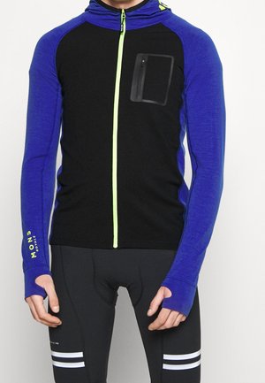 Mężczyzna ubrany w czarno-niebieski, długi rękaw rowerowy z otworami na kciuki i neonowożółtym zamkiem, w połączeniu z czarnymi spodenkami rowerowymi.