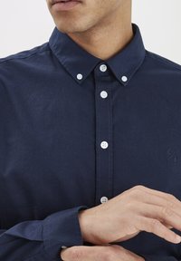 Sötétkék gombos ing sima anyagból, klasszikus gallérral, fehér gombokkal és hímzett logóval a mellkasán.