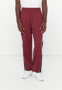 Bordeaux trackbukser med sorte sidelister, elastisk talje og lommer; parret med hvide sneakers. Stoffet virker let og glat.