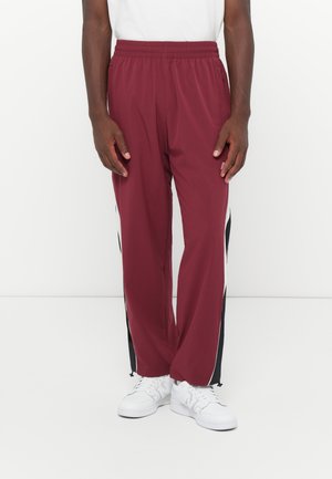 Maroon track pants with black side stripes, elastic waistband, and pockets; paired with white sneakers. Fabric appears lightweight and smooth.