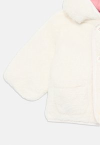 Vit fluffig kappa med en mjuk textur, stor krage och framfickor. Har en rosa foder och en knappstängning framtill.