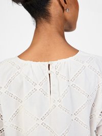 White blouse with intricate cutout diamond pattern, rounded neckline, button closure with pearl accent, and loose fit. Soft, lightweight fabric.