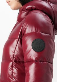 Close-up van een persoon die een glanzende rode pufferjack met capuchon en een zwart ronde patchlogo op de bovenarm draagt.
