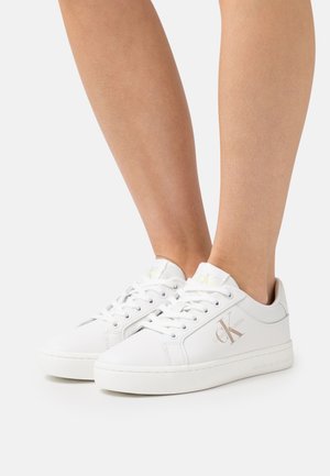 Ein Paar weiße Calvin Klein Low-Top-Sneaker, die barfuß getragen werden, mit seitlichem Logo und geschnürtem Obermaterial vor einem sauberen Hintergrund.