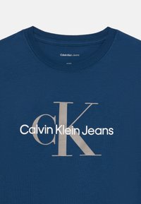 Jūras zila kokvilnas T-krekls ar lielu balto logo "Calvin Klein Jeans", kurā izceltas ievērojama sudraba burts "K" un pelēki akcenti.