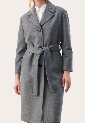 Femme portant un manteau gris en laine longueur genoux avec une ceinture nouée à la taille, trois boutons noirs, et une chemise blanche à col en dessous.