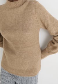 Beżowy sweter z prążkowanego dzianiny z wysokim kołnierzem i długimi rękawami. Materiał wydaje się miękki o nieco grubym splocie, zaprojektowany z myślą o komforcie.
