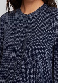 Laivastonsininen pusero, joka on valmistettu pehmeästä kankaasta. Siinä on pyöreä pääntie, lyhyt nappilista ja valkoinen pilkkuinen kuosi kauttaaltaan. Rakenne on teksturoitu.