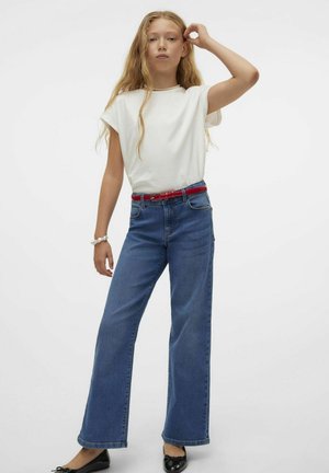 Hvid kortærmet bluse, blå brede jeans med et rødt bælte, sorte sko, langt bølget hår, står imod en ensfarvet hvid baggrund.