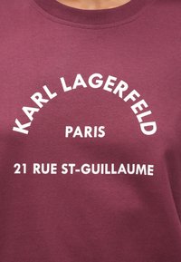 Bordová košeľa s bielym textom "KARL LAGERFELD" zakriveným nadol, "PARIS" umiestneným v strede pod ním a "21 RUE ST-GUILLAUME" na spodnej časti.