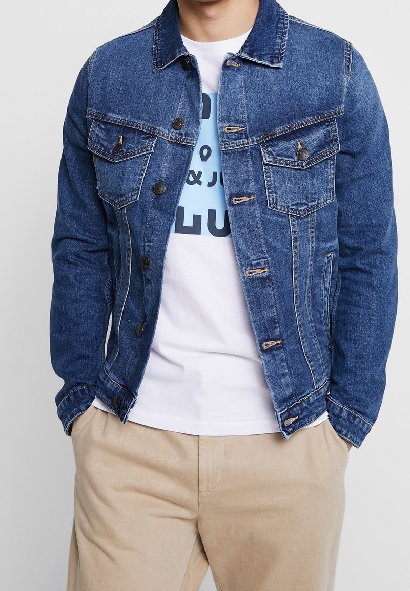 Man wearing a blue denim jacket over a white graphic t-shirt and beige pants, with hands in pockets, against a plain background.