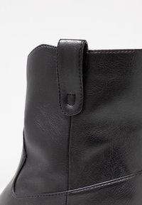 Gros plan sur la partie supérieure d'une botte en cuir noir montrant une languette cousue et la texture détaillée de la surface du cuir.