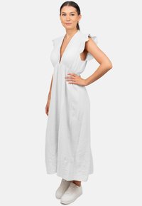 Robe maxi blanche avec des manches volantées courtes, décolleté en V profond, tissu texturé, silhouette fluide, portée avec des baskets blanches.
