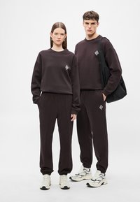 Tmavohnedé zladené mikiny a tepláky s malým vzorovaným logom. Hladká látka, voľný strih, s elastickými manžetami a pásom.