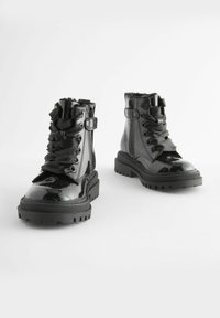 Bottes noires vernies à mi-mollet avec semelles épaisses, larges lacets en satin, zips latéraux et détails texturés sur fond blanc.