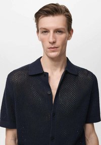 Maglietta a maniche corte in maglia blu navy con un design a trama aperta e colletto. Presenta una chiusura con bottoni e una leggera lucentezza.