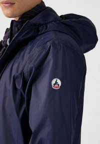 Marineblauwe lichtgewicht jas met een capuchon, met een gladde textuur en een JOTT-logo patch op de linker mouw. Ritssluiting.