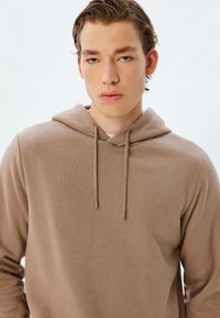 Sudadera de algodón marrón con capucha ajustable, que presenta una textura suave y un corte relajado. No se observan patrones notables ni acentos de hardware.
