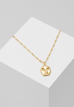 Guldhalskæde med en vedhæng formet som et stiliseret ansigt med hævede øjne og næse, vist på en hvid geometrisk stand.