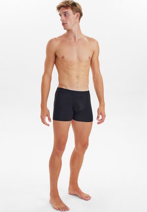 Schwarze Boxershorts aus glattem Stoff, mit einem elastischen Bund und enger Passform, präsentiert von einem männlichen Model vor einem einfarbigen Hintergrund.