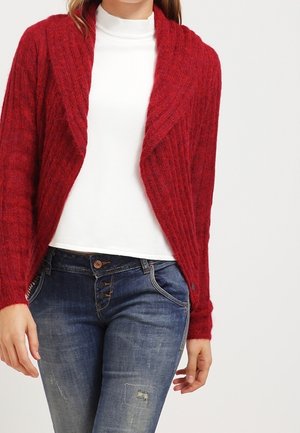 Kvinde iført en rød åben-front strikket cardigan over en hvid rullekrave og blå falmede jeans med knapdetaljer.