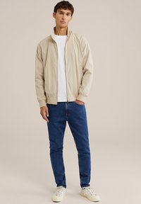 Lahka bež bomber jakna, bela majica in modri jeans slim fit, v kombinaciji z belimi čevlji, proti nevtralni ozadju.