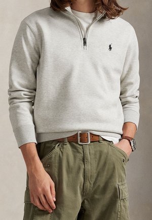 Mann trägt hellgrauen Sweatshirt mit Viertel-Reißverschluss und Logo, olivgrüne Cargohose, braunen Ledergürtel und Armbanduhr, Hand in der Hosentasche.