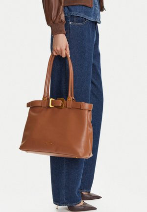 Person trägt blaue Jeans und braune Jacke und hält eine große braune Ledertasche mit goldener Schnalle und zwei Griffen.