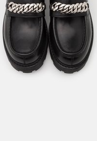 Musta nahkakengät, joissa on paksut pohjat ja tukevat hopeaketju yksityiskohtina päällä, kuvattuna ylhäältä valkoista taustaa vasten.