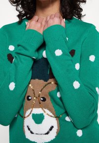 Pull en maille verte avec un motif de renne portant des lunettes à sequins, un motif à pois et des poignets côtelés. Texture douce et coupe décontractée.