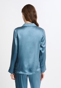 Parte superior de pijama de satén en color teal, con cuello clásico, mangas largas y textura suave, mostrando la vista trasera.