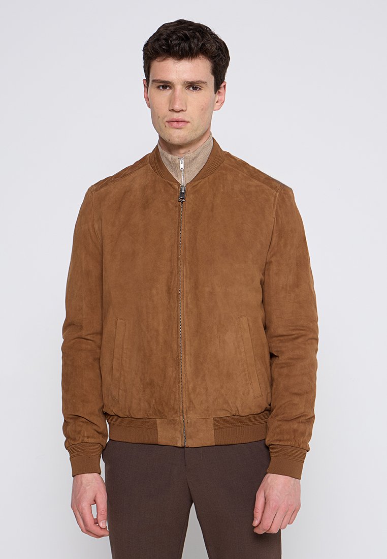 Młody mężczyzna w brązowej zamszowej kurtce bombowcu z ściągaczami przy mankietach i na dole, stojący na tle jasnego, jednolitego tła.