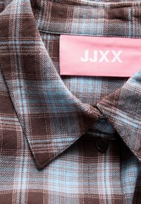 Gros plan sur le col d'une chemise en tissu à carreaux marron et bleu avec une étiquette rose affichant les lettres blanches JJXX.