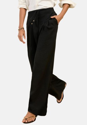 Femeie care poartă pantaloni negri largi cu șnur și buzunare laterale, asortați cu o cămașă albă cu mâneci suflecate și sandale negre.