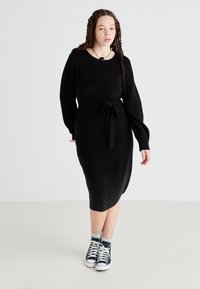 Vestido de punto negro con mangas largas y un cinturón en la cintura, con escote redondo y longitud hasta la mitad de la pantorrilla, combinado con zapatillas negras.