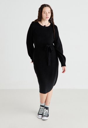 Black knitted dress with long sleeves and a belt at the waist, featuring a round neckline and a mid-calf length, paired with black sneakers.