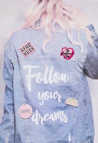 Chaqueta de mezclilla con texto manuscrito en blanco "Sigue tus sueños", adornada con cuatro pines coloridos: "Amabilidad", "Amor", un diseño de corazón, y "Mantente fresco".