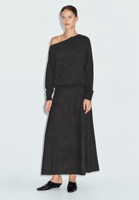 Dunkelgrauer, schulterfreier Strickpullover, kombiniert mit einem passenden, figurbetonten langen Rock. Das Modell trägt schwarze Slippers. Glatte Textur, minimalistisches Design.