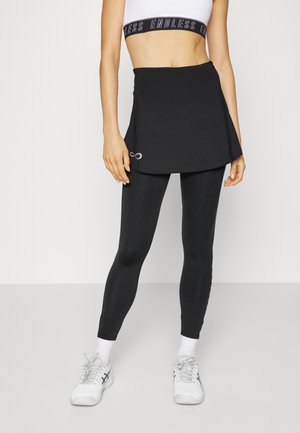 Kobieta ubrana w czarne legginsy z dołączoną spódniczką, biały stanik sportowy z paskiem "ENDLESS", białe skarpetki i białe buty sportowe, stojąca.